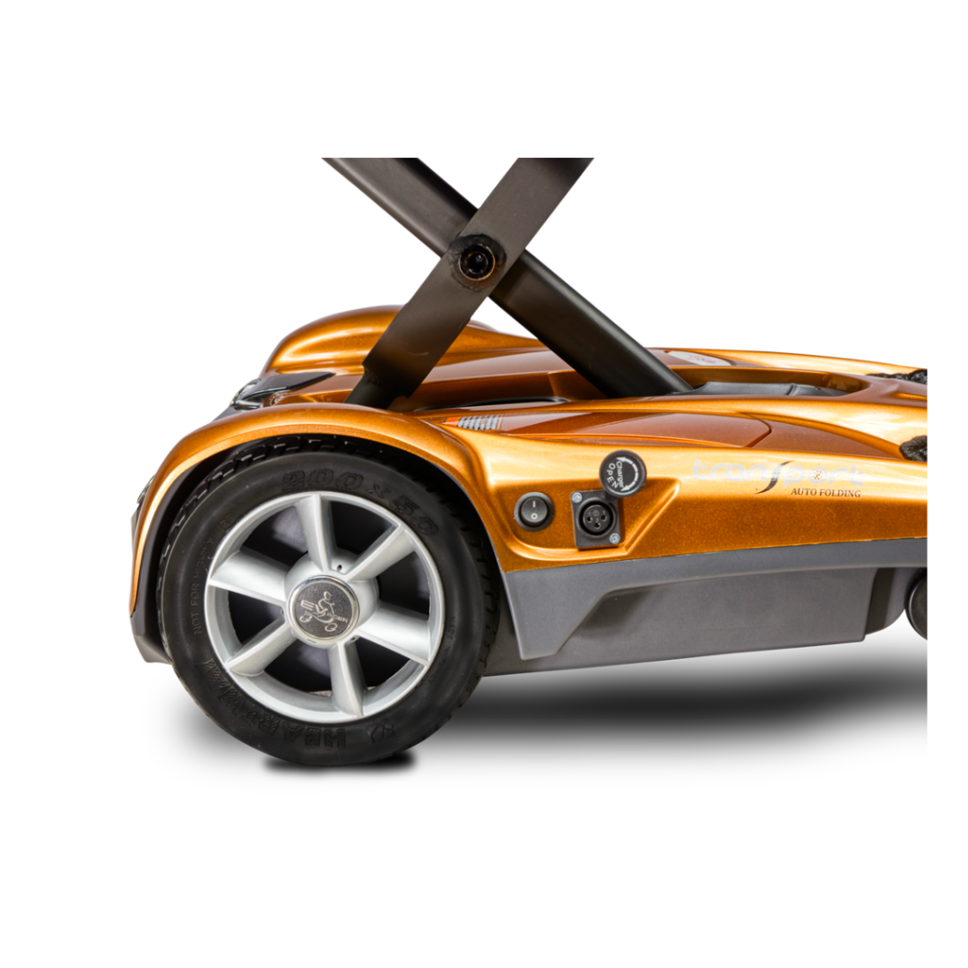 Close-up of the rear wheel and lower body of the EV Rider TranSport S19AF Automatic Folding Travel Scooter in orange and black, highlighting part of the footboard, wheel design, and charging port against a white background.