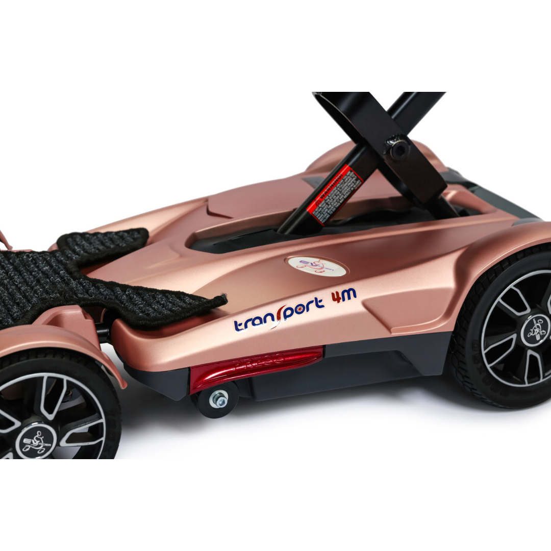 Close-up of the EV Rider Transport 4M Travel Scooter’s rose gold footrest and wheel, featuring the 4M logo, part of the frame, a black textured footplate, and detailed wheel design.