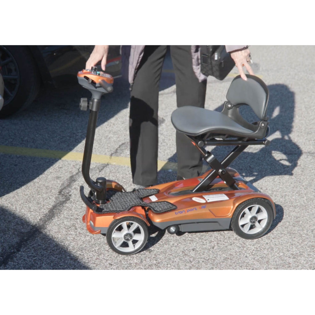 A person stands next to the EV Rider Transport AF4W Automatic Folding Mobility Scooter, an orange compact scooter with a raised seat and handlebars in a parking lot—ideal for convenient, portable mobility and easy transport.