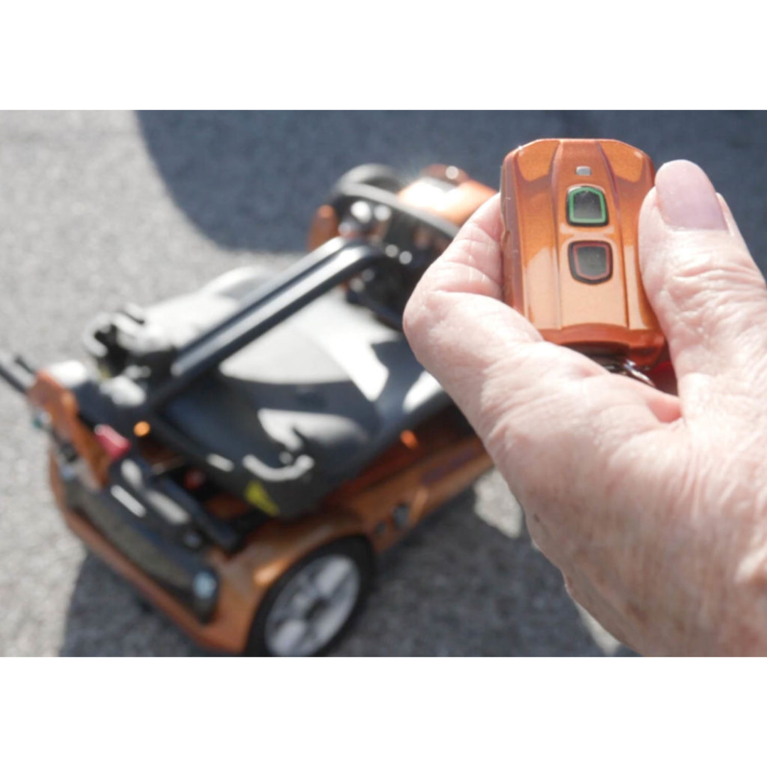A hand holds a small orange remote with green and red buttons, aimed at the EV Rider Transport AF4W Automatic Folding Mobility Scooter on a sunny outdoor surface, ideal for easy transport and convenience.