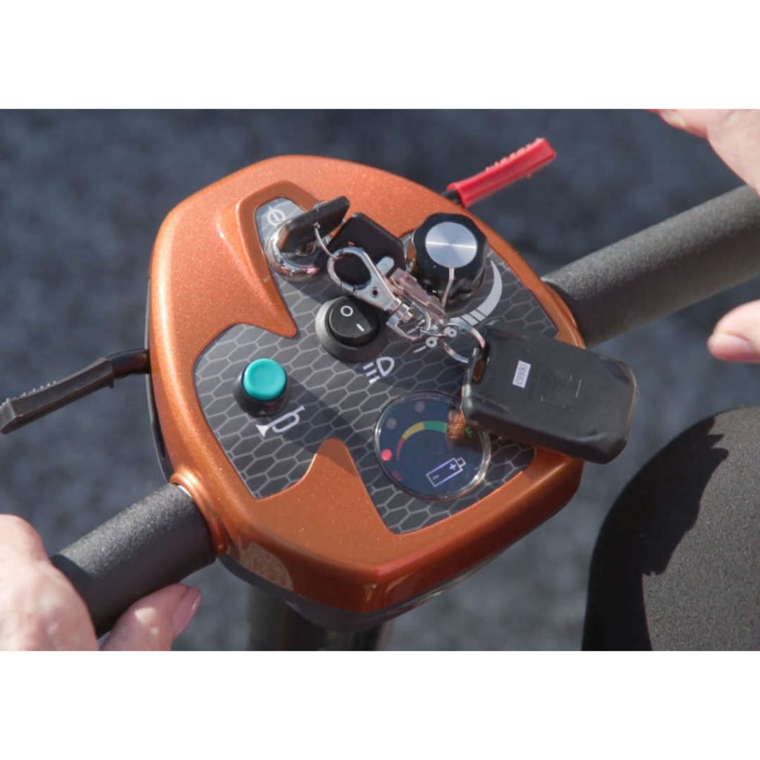 Close-up of hands operating the EV Rider Transport AF4W Automatic Folding Mobility Scooter control panel, featuring buttons, a key, a dial, and a gauge display on its textured orange console.
