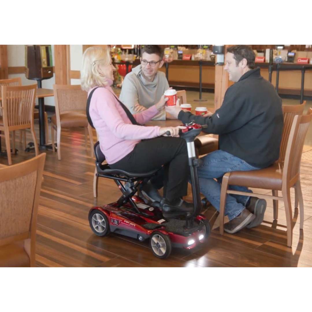 A woman on an EV Rider Transport AF4W Automatic Folding Mobility Scooter sits at a wooden table with two men in a casual dining area, all smiling and engaged in conversation.