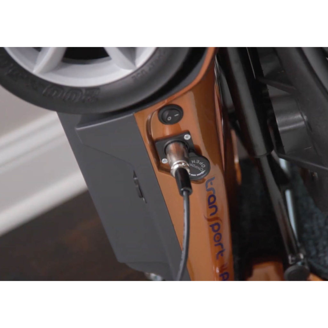 Close-up of the EV Rider Transport AF4W Automatic Folding Mobility Scooter being charged, with the cable connected to the port on its orange frame. A wheel appears at the top of the image.