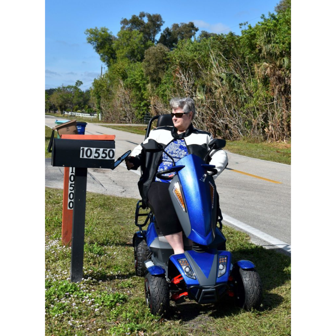 An older woman in sunglasses sits on a blue Heartway S12X Vita Monster All-Terrain Power Scooter, reaching into a roadside mailbox marked 10550 on a sunny day, with trees and a quiet road in the background.