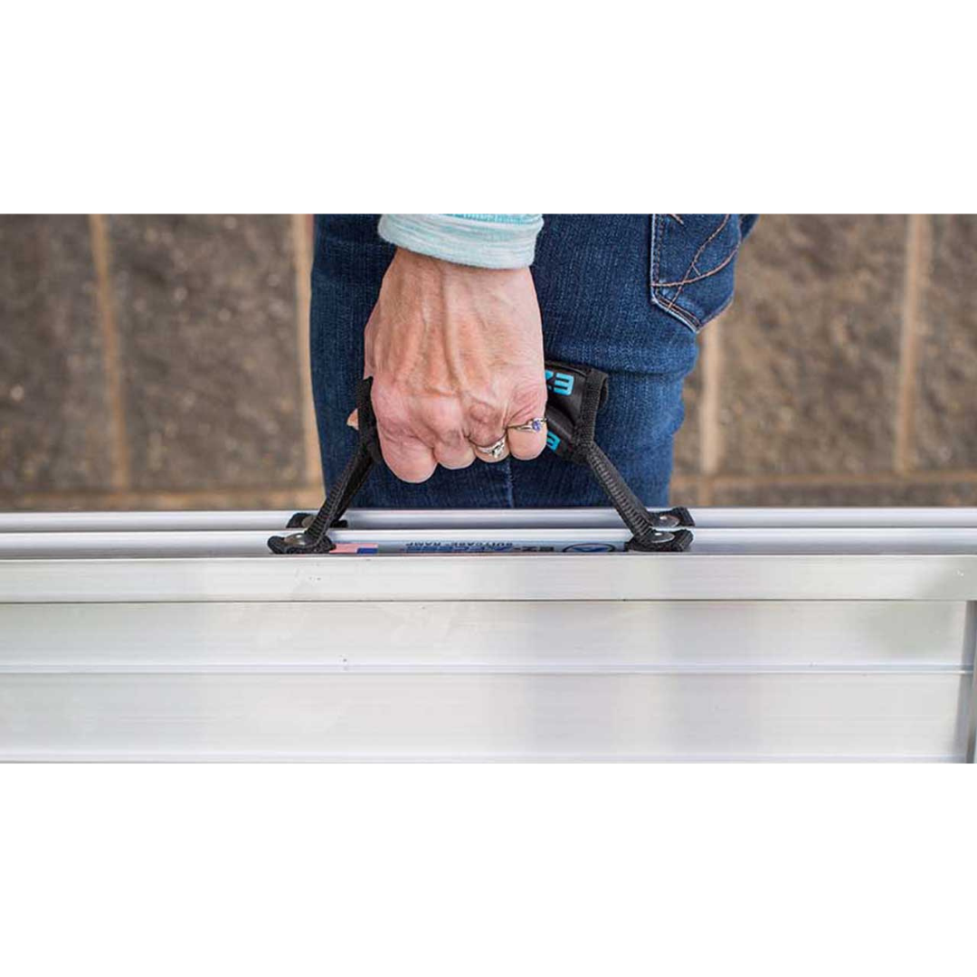 A person wearing jeans stands before a brick wall, holding the black handle of an EZ-Access SUITCASE® Singlefold AS Portable Ramp with a slip-resistant surface.