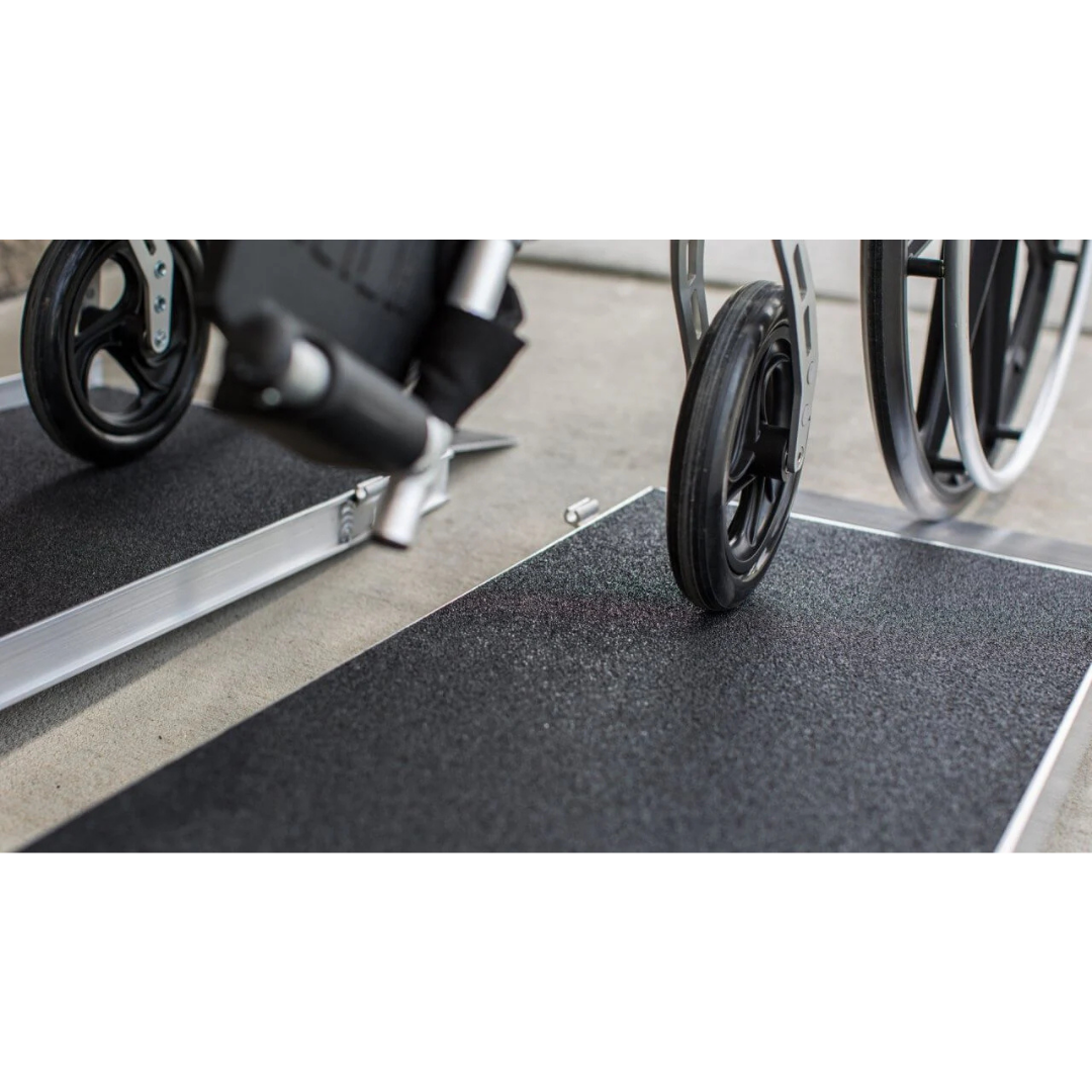 Close-up of a wheelchair wheel moving up an EZ-Access SUITCASE® Singlefold AS Portable Ramp with a slip-resistant surface on a concrete floor.
