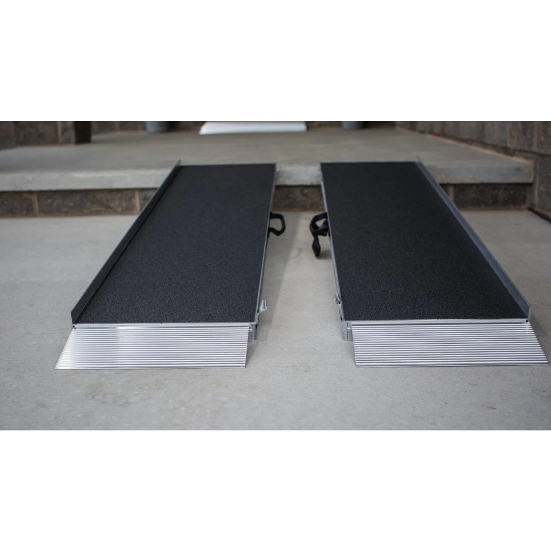 Two EZ-Access SUITCASE® Singlefold AS Portable Ramps with slip-resistant surfaces and metal edges are positioned side by side, providing access up a concrete step at a doorway.