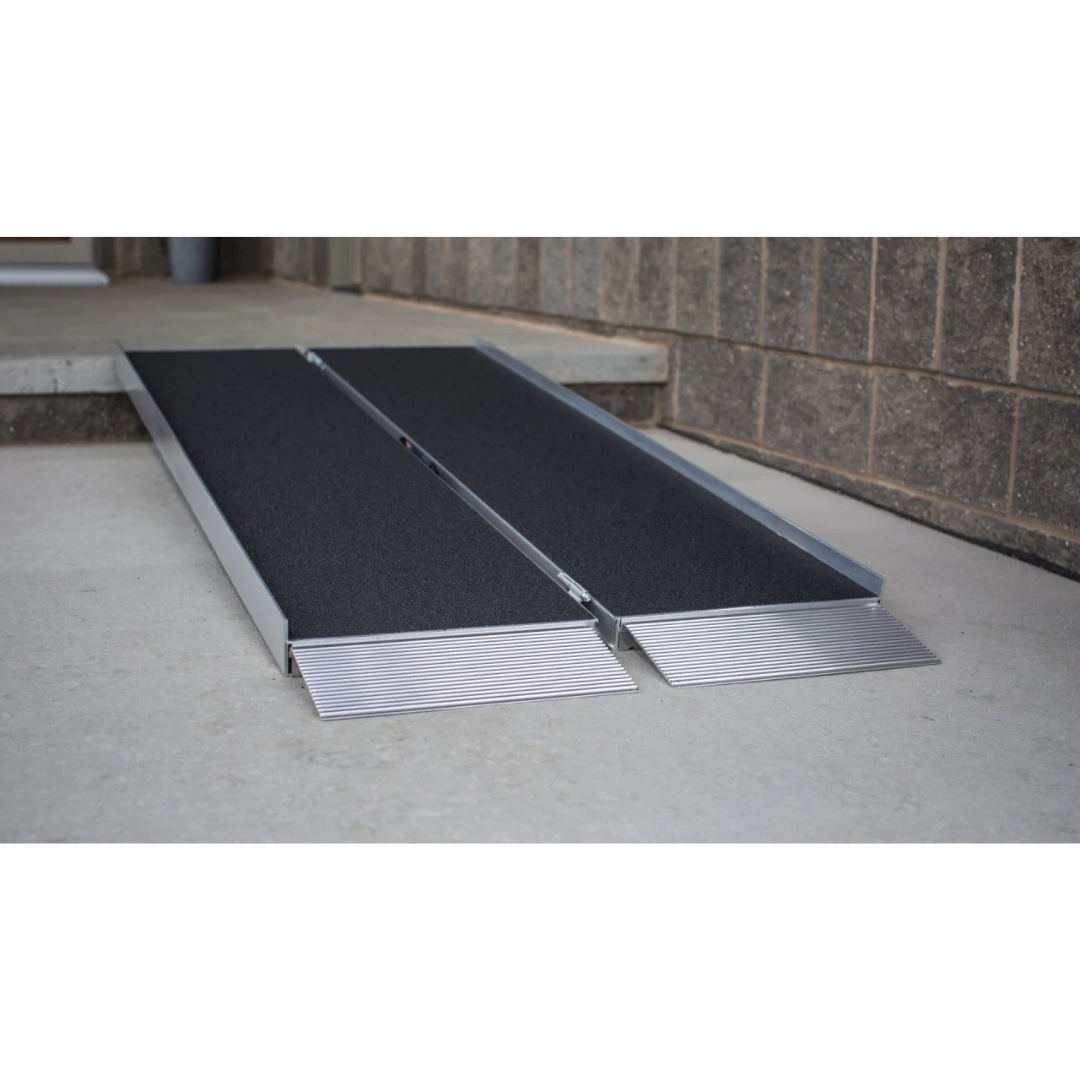An EZ-Access SUITCASE® Singlefold AS Portable Ramp with a slip-resistant surface bridges a small step, connecting a concrete sidewalk to a raised entrance beside a brick wall.