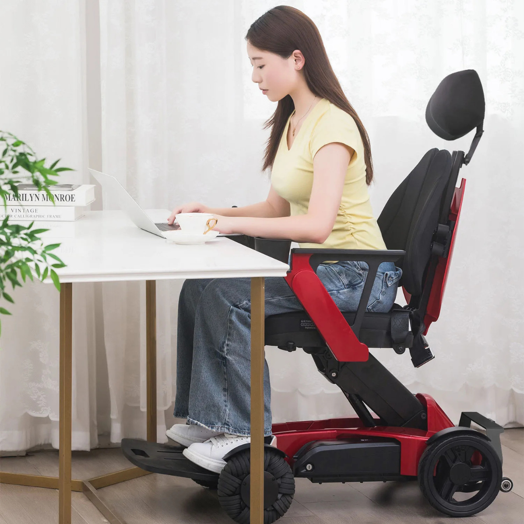 A woman in a yellow shirt and jeans sits indoors at a white desk using a laptop on the Easwe™ L10 Luxury Electric Wheelchair with Automatic Adjustable Seat Height, with plants and framed art visible in the background.