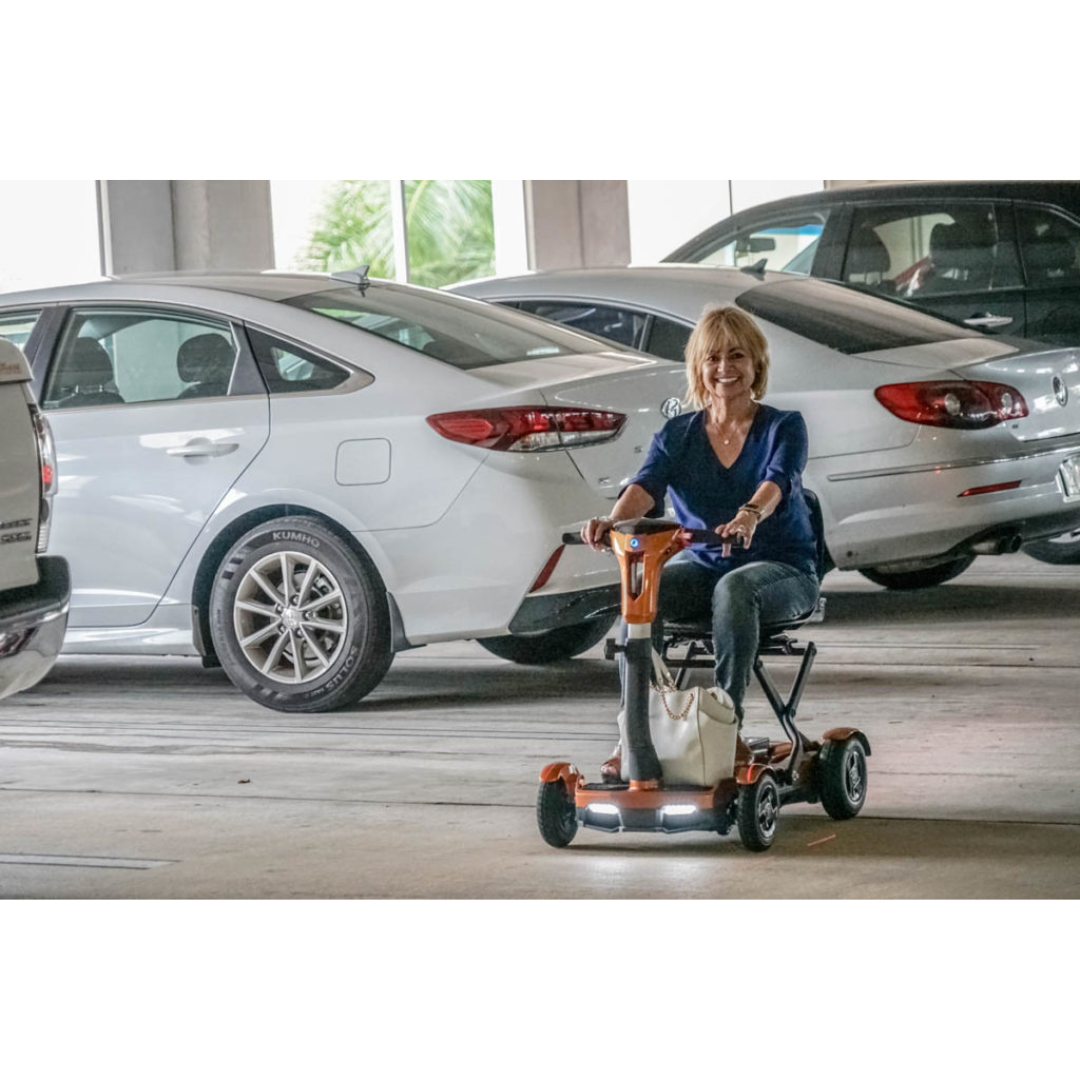 A smiling woman rides an EV Rider Teqno Automatic Folding Portable Travel Scooter through a parking garage, passing parked cars. She wears a blue top and jeans, with a white bag hanging from the scooter as daylight streams in behind her.