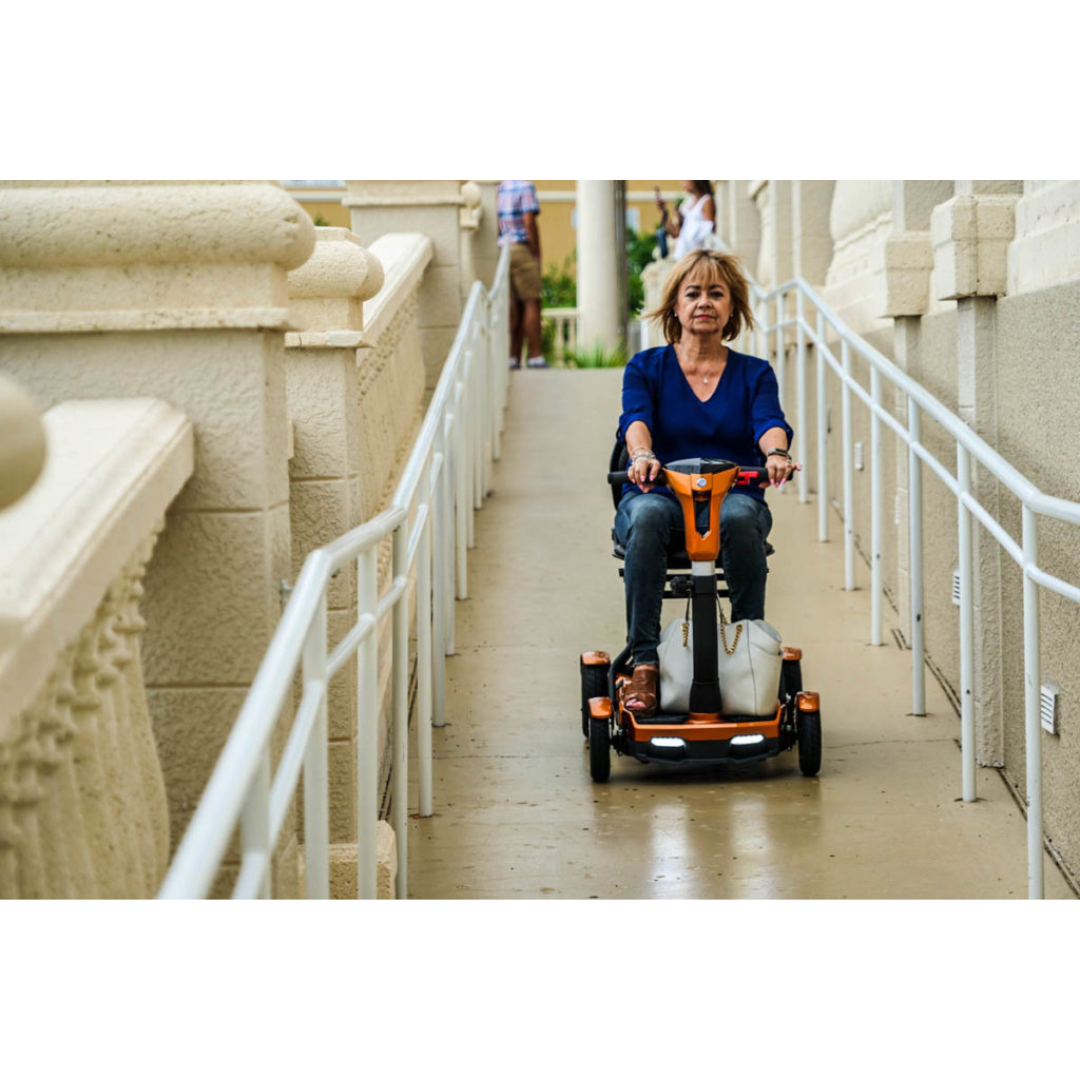 A woman rides the EV Rider Teqno Automatic Folding Portable Travel Scooter down an accessible ramp with railings, surrounded by beige stone walls. She is focused, wearing a blue top and jeans.
