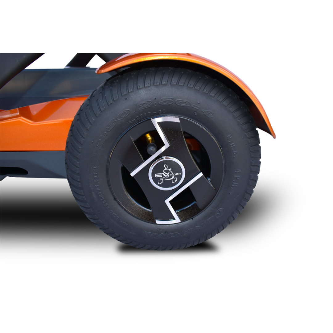 Close-up of a black rubber tire with visible treads and a black and silver rim featuring a stylized cyclist emblem, attached to an orange EV Rider Teqno Automatic Folding Portable Travel Scooter.