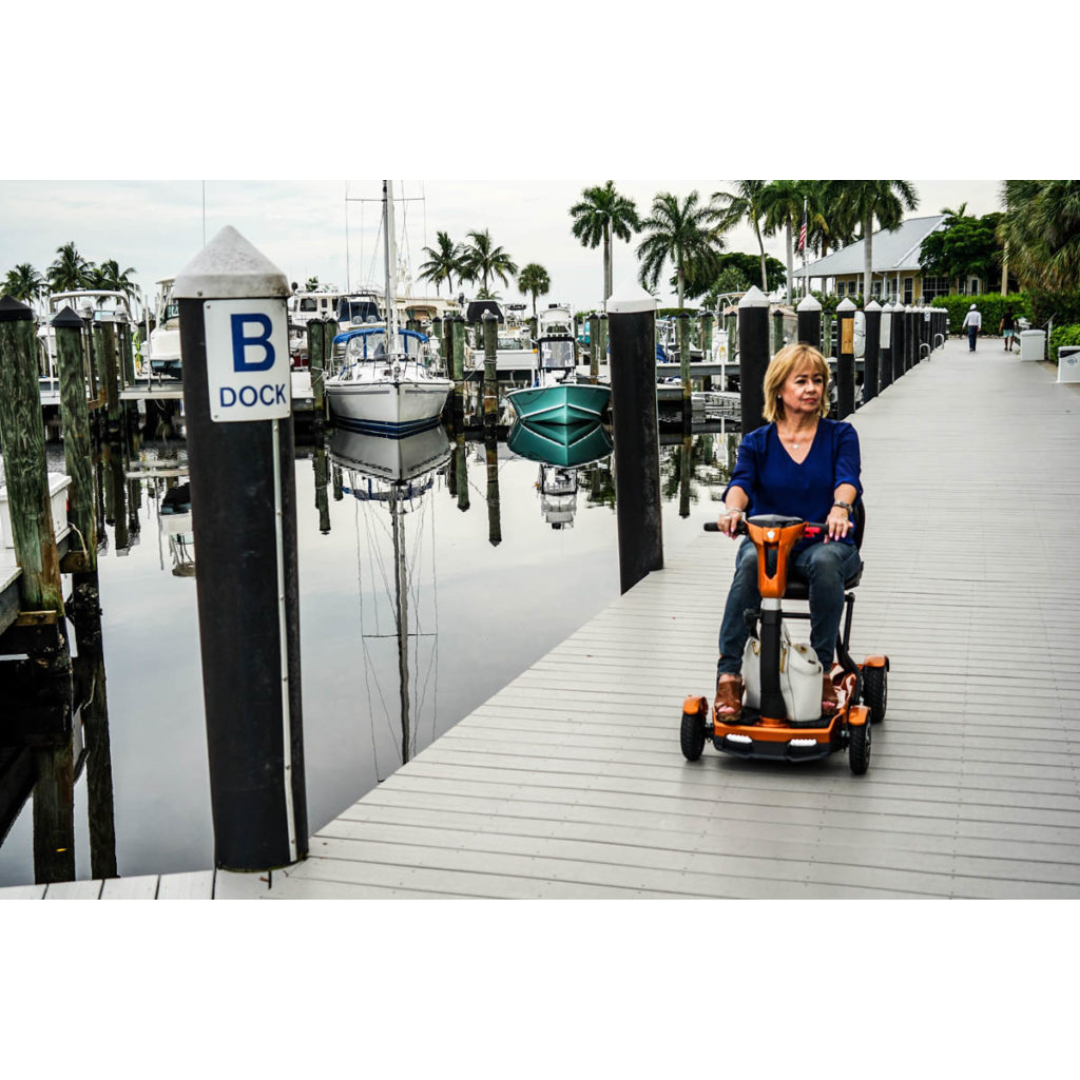 A woman rides an EV Rider Teqno Automatic Folding Portable Travel Scooter on a wooden dock near a marina with boats and palm trees, beside a "B Dock" sign. The calm scene features partly cloudy skies.