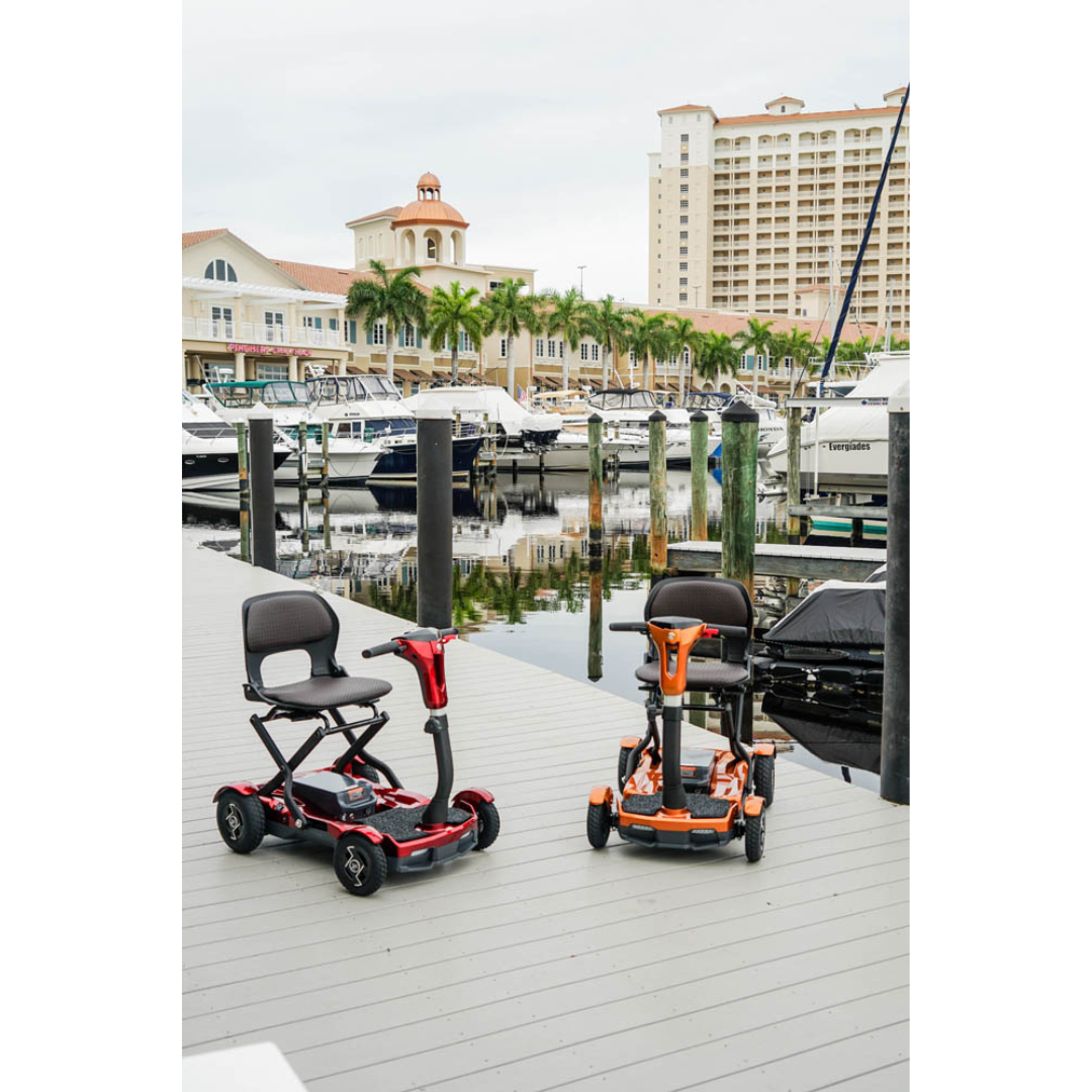 Two EV Rider Teqno Automatic Folding Portable Travel Scooters, one red and one orange, are parked on a dock beside a marina with boats, with palm trees and tall buildings visible under a cloudy sky.