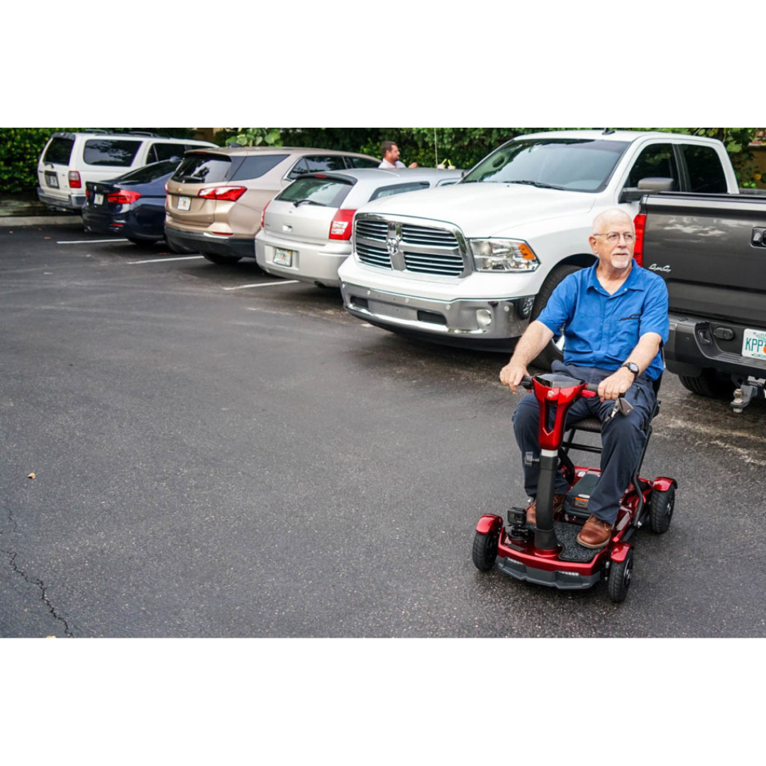 An older man in a blue shirt rides an EV Rider Teqno Automatic Folding Portable Travel Scooter through a parking lot, passing parked cars and trucks, with trees and another person visible in the background.