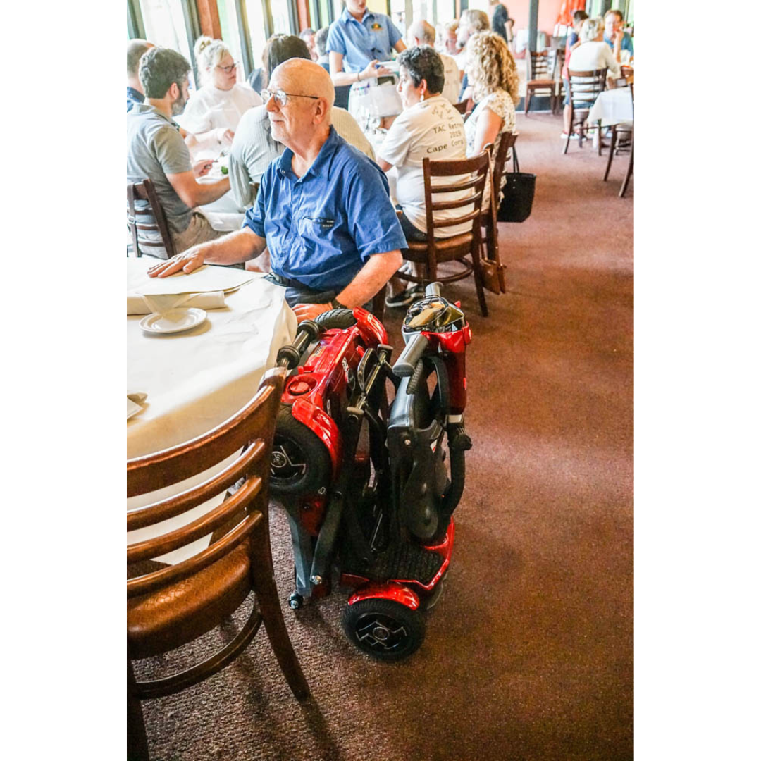 An older man with glasses sits at a restaurant table among other diners. Next to him is an EV Rider Teqno Automatic Folding Portable Travel Scooter. The room is filled with people seated at tables.