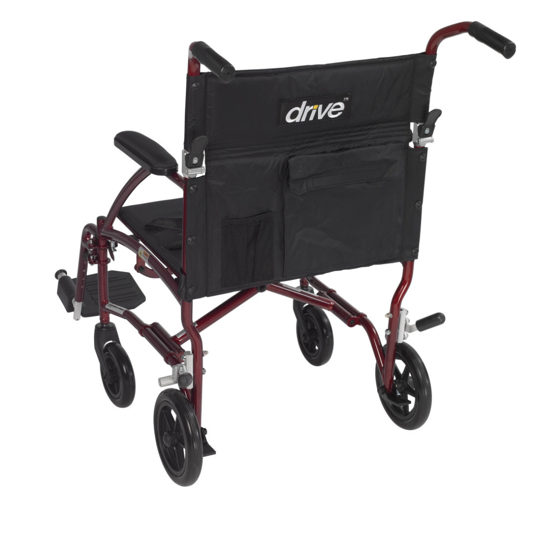 The Drive Fly-Lite Aluminum Transport Chair features a lightweight, foldable metallic red frame, black seat and backrest, armrests, footrests, small black wheels, and a rear storage pocket labeled "drive.
