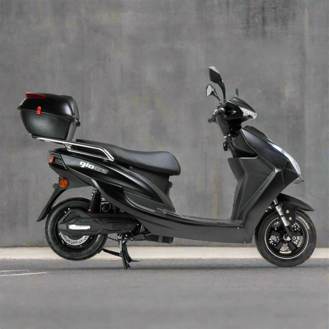 The GIO Phoenix Premium Electric Scooter, an eco-friendly black e-scooter with a rear cargo box, is parked on gray asphalt beside a plain concrete wall.