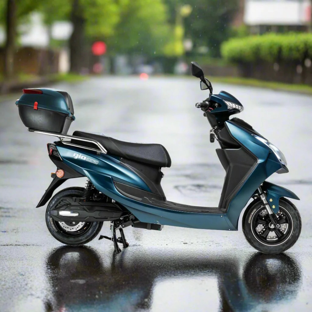 A GIO Phoenix Premium Electric Scooter in blue, featuring a rear storage box, is parked on a wet, empty street bordered by greenery during a rainy day.