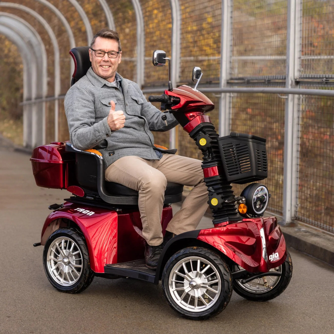 A smiling man in glasses and a gray jacket gives a thumbs-up while sitting on a red GIO Tron Pro 4-wheel mobility scooter beside a metal fence and autumn leaves.