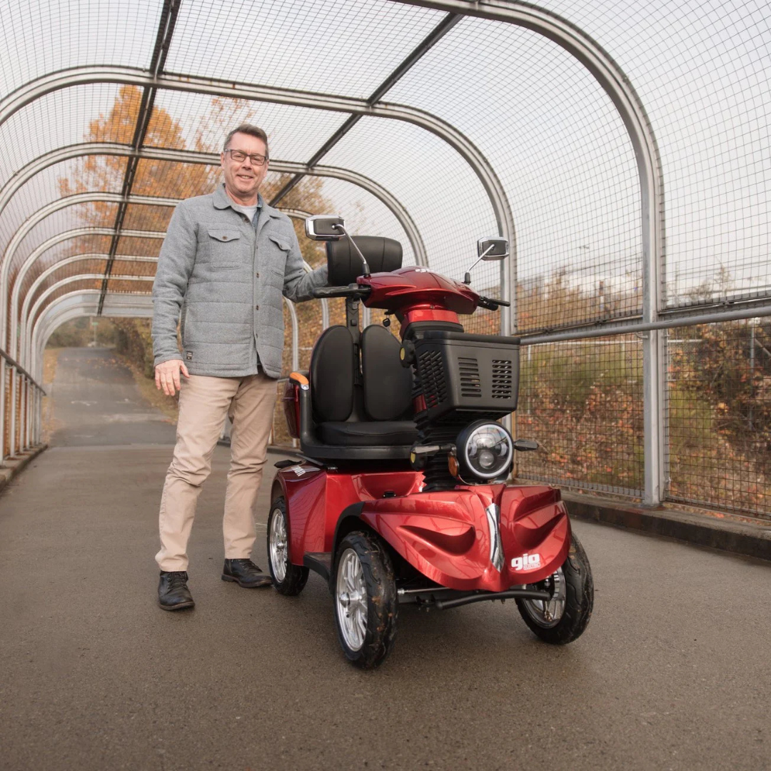 A man in a light gray jacket stands on a covered outdoor path, smiling next to a red GIO Tron Pro mobility scooter with 4-wheel stability. Autumn trees with orange leaves are seen in the background.