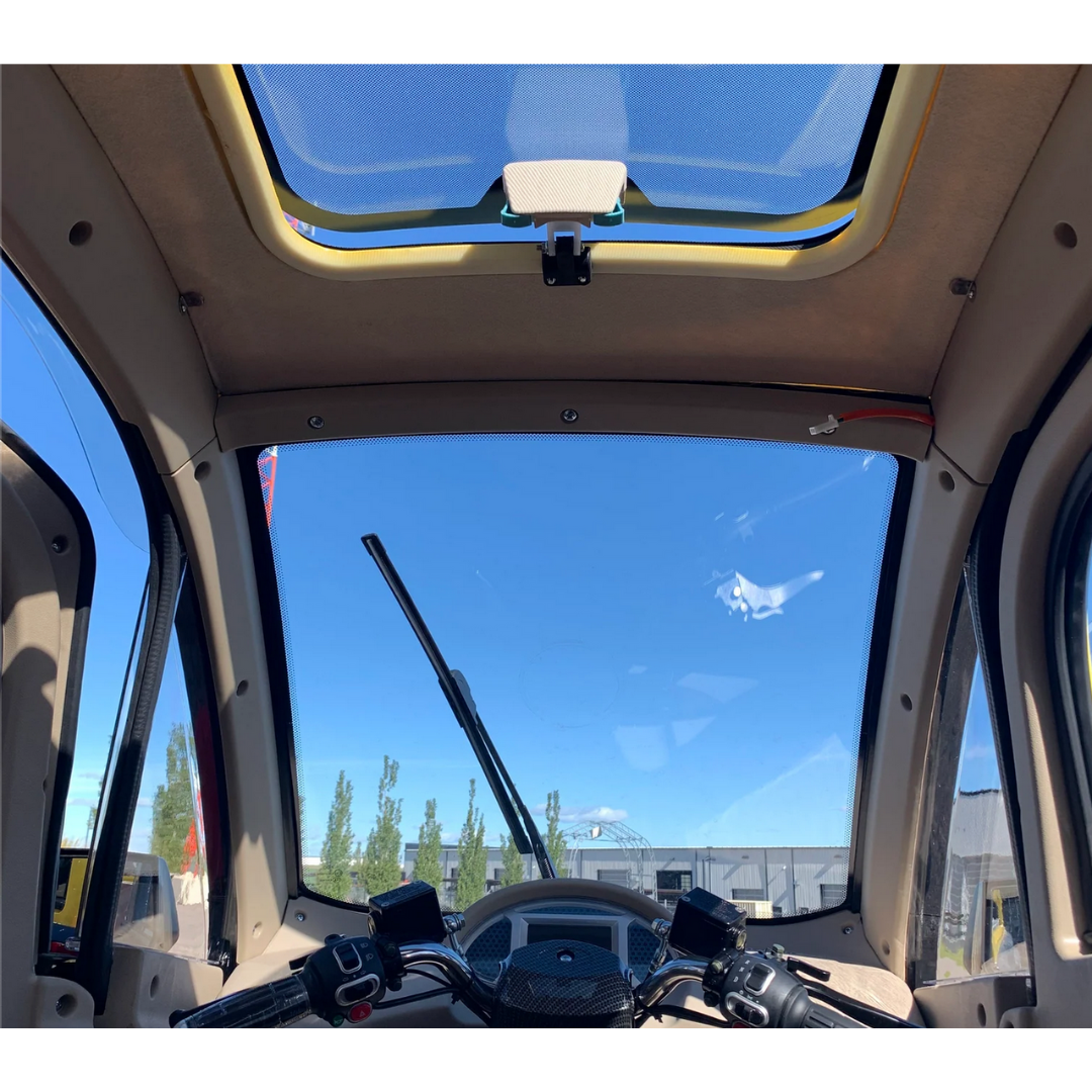 Interior of the GIO Golf Fully Enclosed Mobility Scooter features a clear windshield and sunroof showcasing a blue sky. The dashboard includes navigation and controls. Outside, trees and buildings are visible under the bright sky, providing an enjoyable ride with its heater and cooling fan.