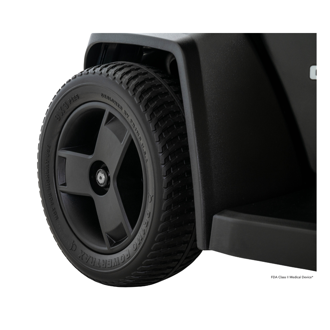 Close-up of a black, textured wheel with tread and embossed sidewall text on the Pride Mobility Go Chair® MED Portable Powerchair, featuring a sturdy plastic fender and platform—ideal for lightweight travel.
