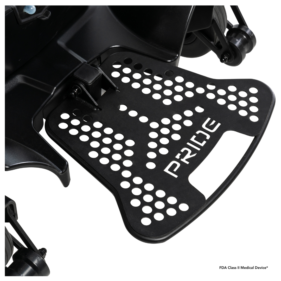 Close-up of a black metal footrest with circular holes and "PRIDE" cut out, attached to the Pride Mobility Go Chair® MED Portable Powerchair; wheels and part of the frame are visible in the background.