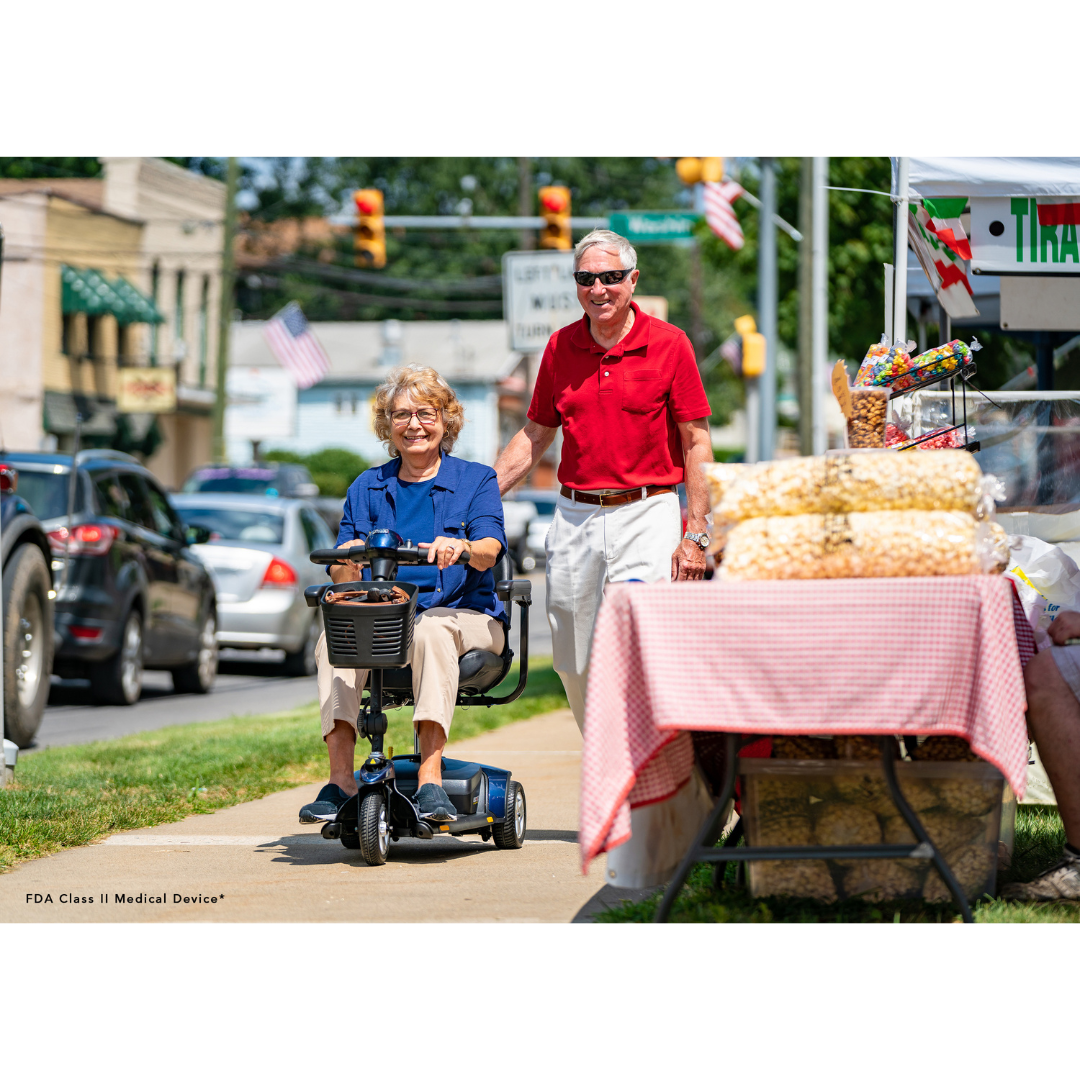 An older woman smiles as she rides her Pride Mobility Go Go® Elite Traveller 3-Wheel Scooter beside a man in a red shirt on a sunny day. They pass a snack stall, and an American flag waves in the background, highlighting the small-town charm.