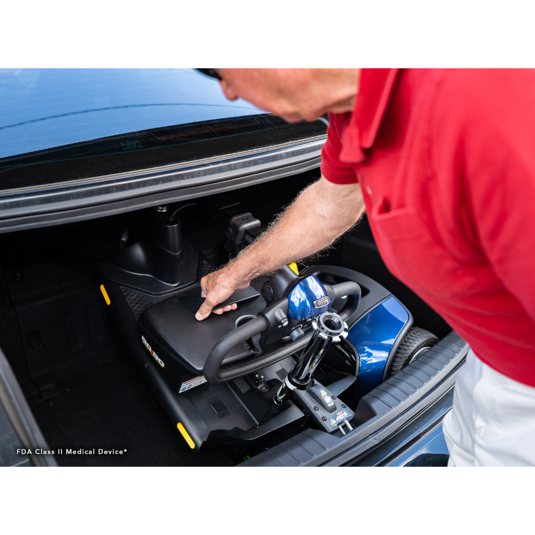 Wearing a red shirt, a person places the Pride Mobility Go Go® Elite Traveller 3-Wheel Mobility Scooter, known for its lightweight and compact design, in the car trunk, showcasing its easy transportability.