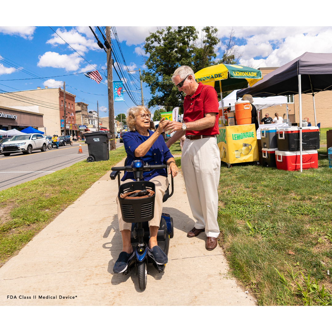 An elderly woman sits on a Pride Mobility Go Go® Elite Traveller 3-Wheel Scooter, smiling with a drink from an elderly man beside her. They enjoy the bustling street fair near a lemonade stand as buildings and tents provide a charming backdrop to their cheerful day outdoors.