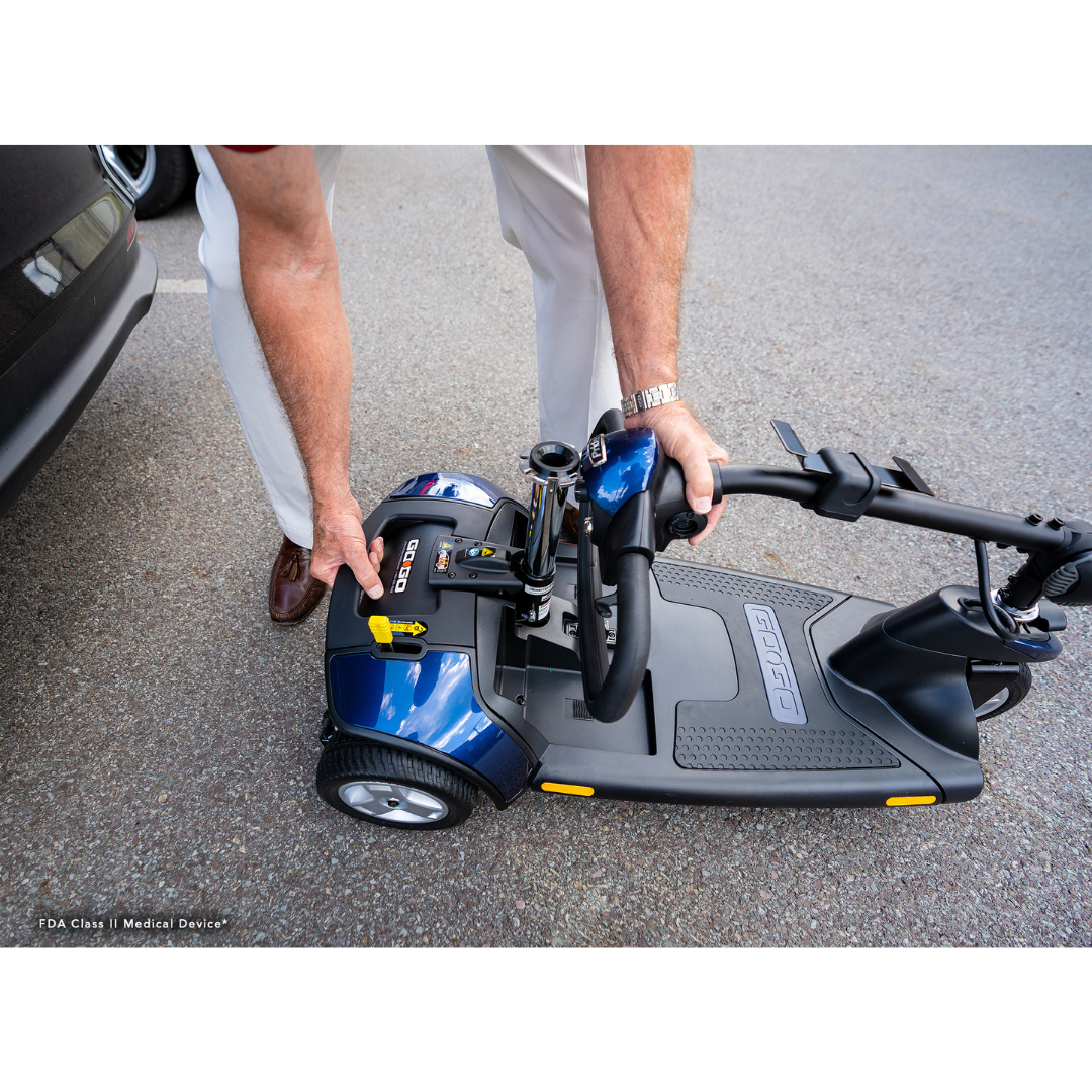 Effortlessly folding a blue Pride Mobility Go Go® Elite Traveller 3-Wheel Mobility Scooter next to a car, the person uses one-handed frame separation. Its compact design features black handlebars and small wheels, emphasizing its lightweight build and portability.