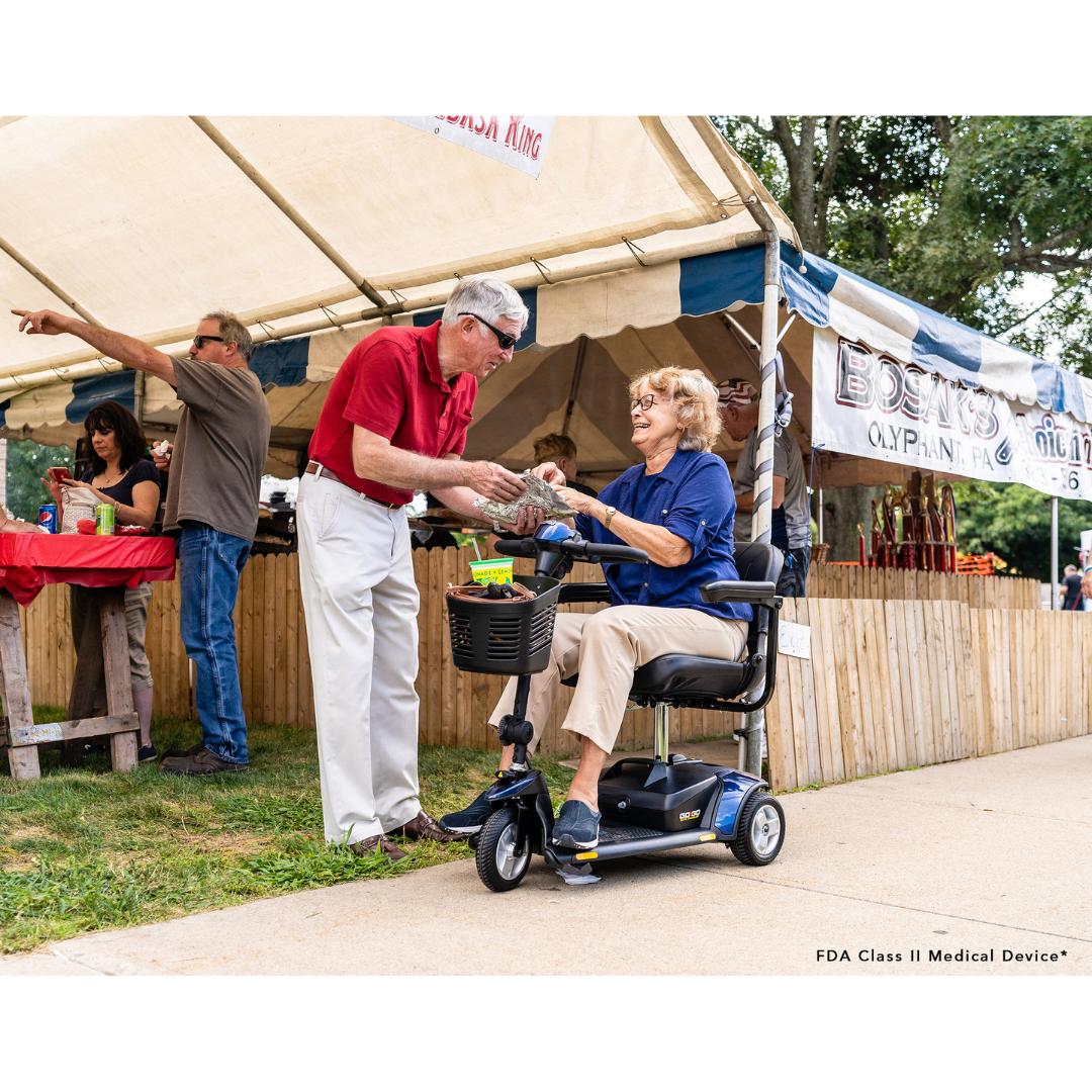 At the fair, a man hands food to a woman on a Pride Mobility Go Go® Elite Traveller 3-Wheel Scooter. She smiles with her sandwich as people gather near a tent with an Olympic sign and food displays.