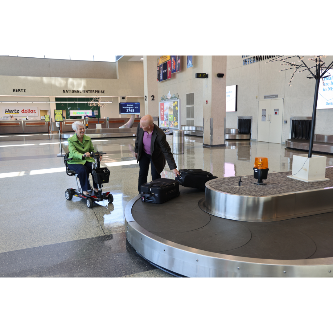 An elderly woman on a Golden Technologies Portable Buzzaround GB120 Carry-On Travel Scooter and an elderly man stand near a baggage carousel in an airport. The man is picking up luggage from the carousel, while car rental counters are visible in the background.