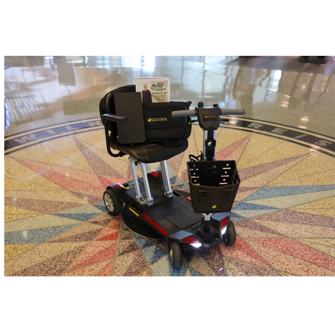 A Golden Technologies Portable Buzzaround GB120 Carry-On Travel Scooter is parked on a terrazzo floor with a compass design. It features a padded seat, backrest, and front basket. A sign labeled EZ-Fold adorns the handle, suggesting its ultra-portable nature and advanced lithium-ion battery.