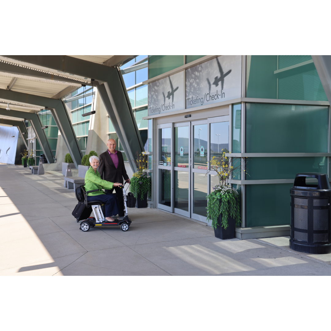 An older woman on a Golden Technologies Portable Buzzaround GB120 Carry-On Travel Scooter and a man stand outside an airport terminal entrance labeled Traveling/Check-In. The modern terminal facade with large glass windows gleams under the sky, while her ultra-portable ride boasts a reliable lithium-ion battery for seamless travel.