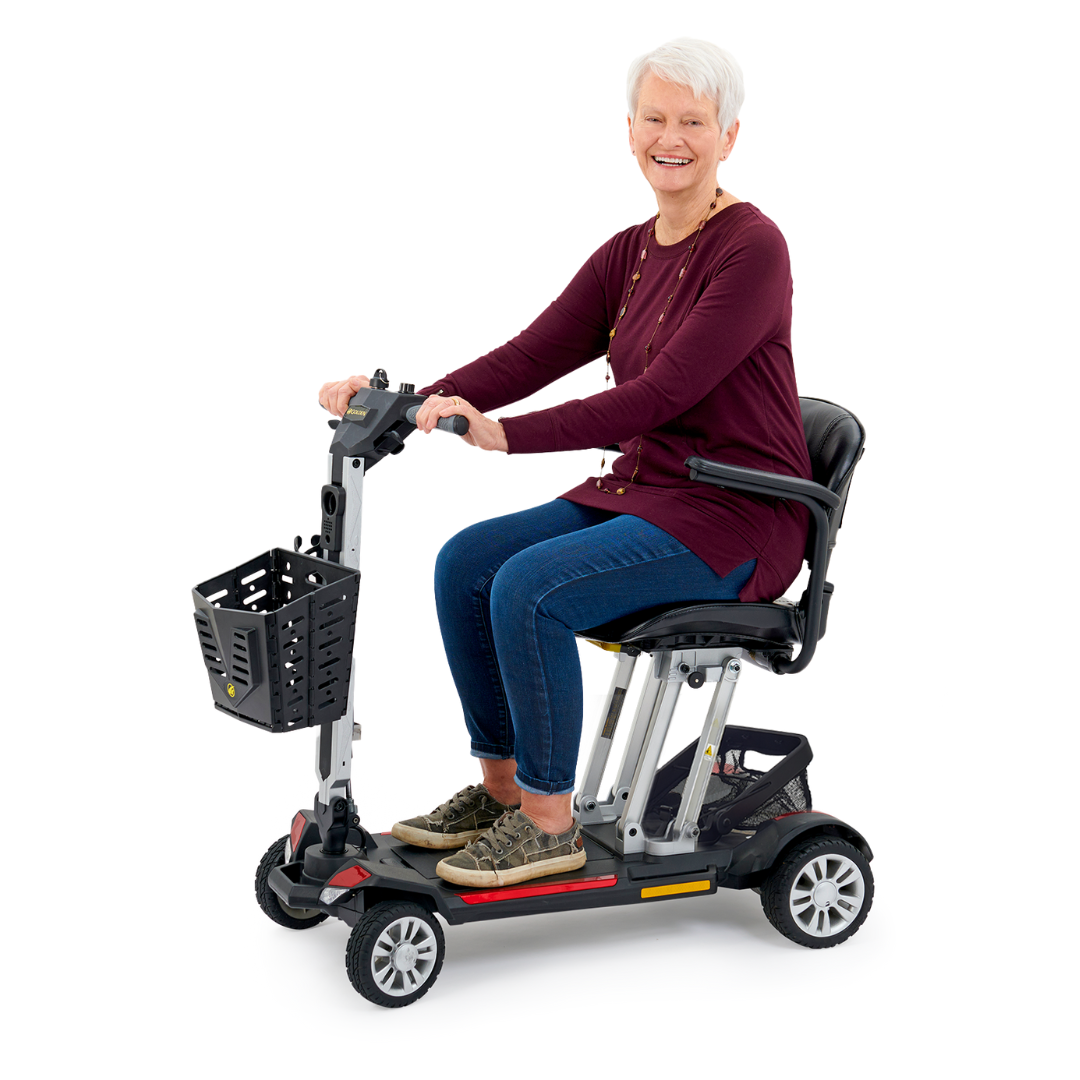 An older adult with short white hair is sitting on the Golden Technologies Portable Buzzaround GB120 Carry-On Travel Scooter, smiling. They are wearing a maroon long-sleeve top and blue jeans. The scooter, equipped with a compact lithium-ion battery, features a small basket in front.
