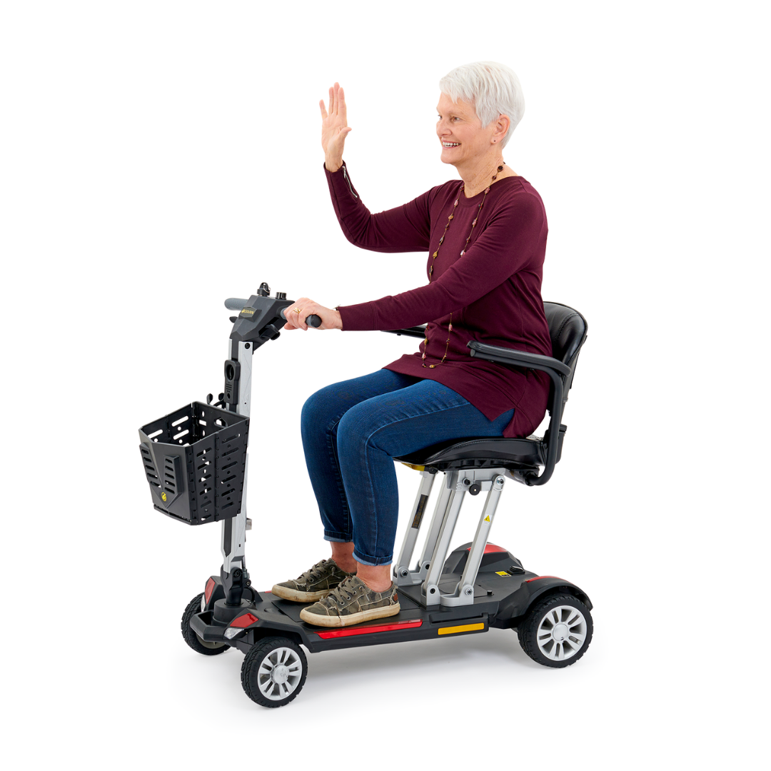 An elderly woman with short white hair is smiling and waving while sitting on the Golden Technologies Portable Buzzaround GB120 Carry-On Travel Scooter. She is wearing a maroon top, blue jeans, and sneakers. This ultra-portable scooter includes a black basket in front and is powered by a long-lasting lithium-ion battery.