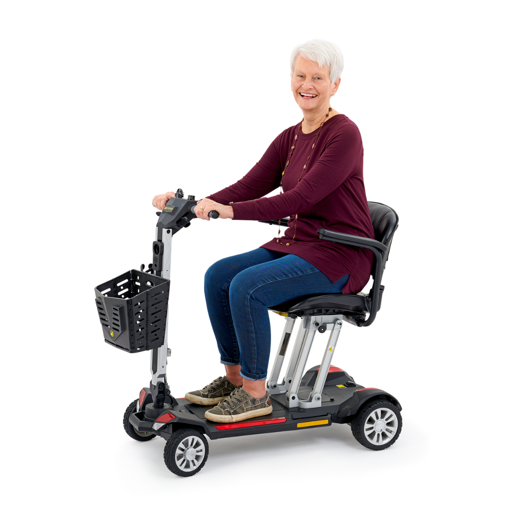 An older adult with short white hair is smiling while seated on a Golden Technologies Portable Buzzaround GB120 Carry-On Travel Scooter. This ultra-portable scooter, powered by a lithium-ion battery, boasts a black basket at the front. The individual is dressed in a dark red top and jeans, perfectly embodying comfort and convenience in motion.