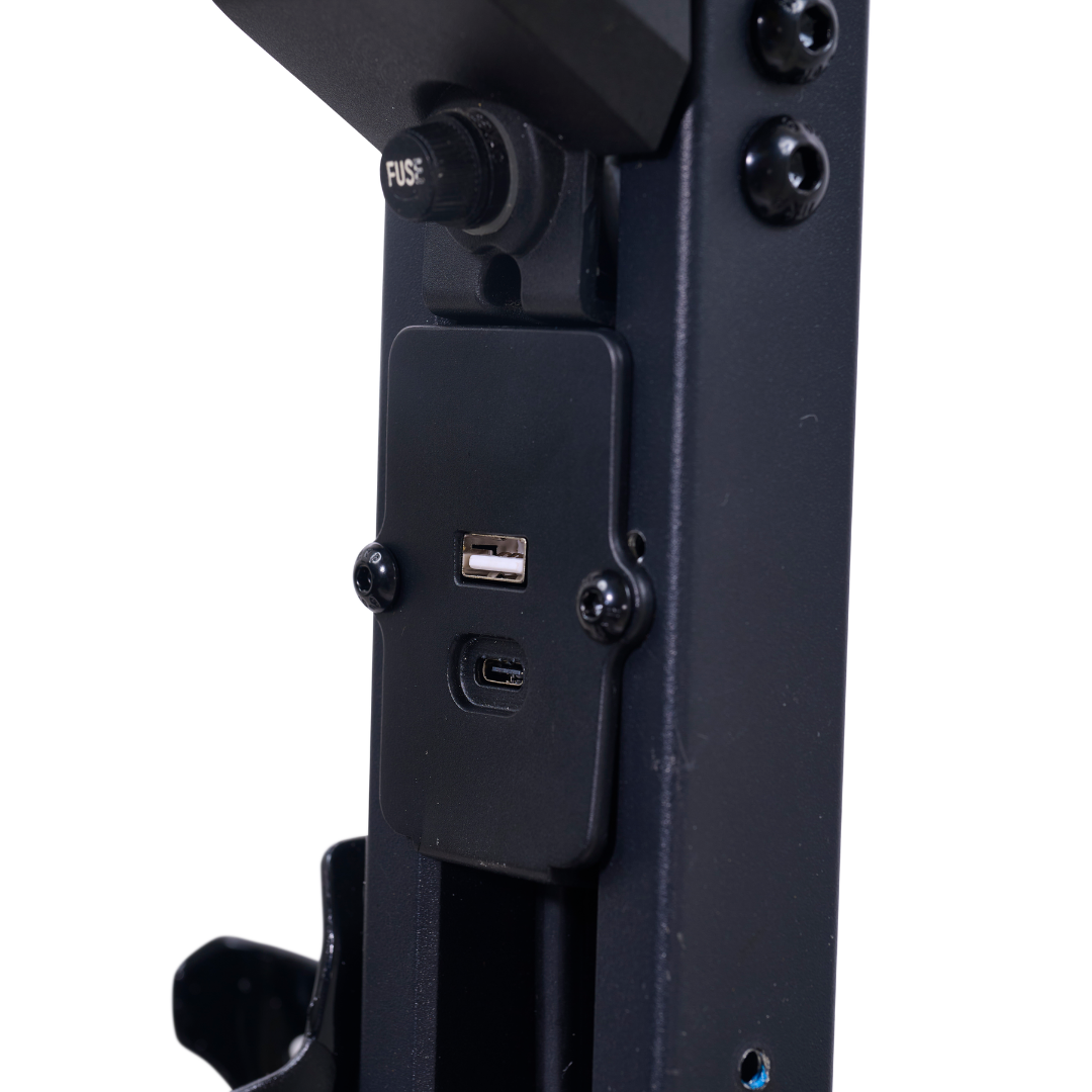 Close-up of a black metal device on the Golden Buzzaround GB220 CarryOn HD Travel Scooter, showing a USB-A port and USB-C port on a rectangular panel, with a fuse button and screws visible above and around the ports.