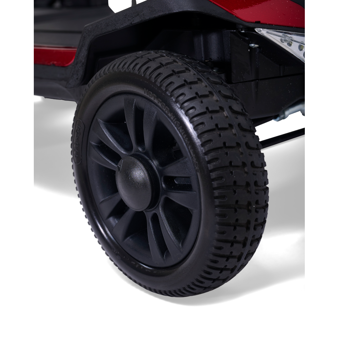 Close-up of a black plastic wheel with detailed tread on the Golden Buzzaround GB220 CarryOn HD Travel Scooter, shown in red and black on a white background.
