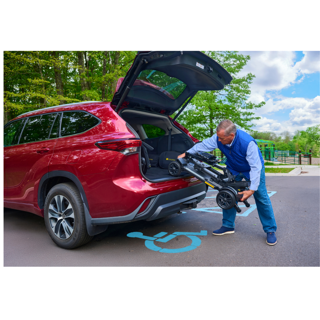 A man loads the folded Golden Buzzaround GB220 CarryOn HD Travel Scooter into the trunk of a red SUV parked in a handicap space on a sunny day, with green trees and blue sky in the background.