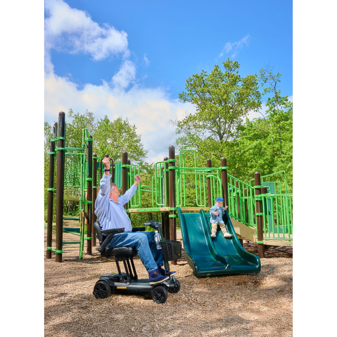 An older man on a Golden Buzzaround GB220 CarryOn HD Travel Scooter raises his arms joyfully beside a young child at the bottom of a green playground slide, all surrounded by trees on a sunny day.
