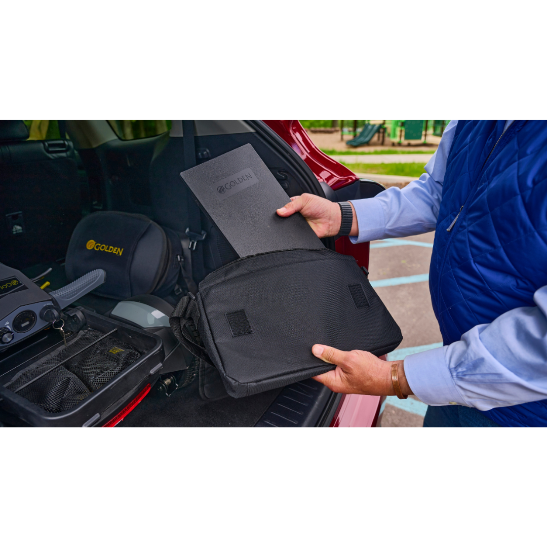 A person puts a black rectangular pad labeled GOLDEN into a black bag in the open trunk of a red car, next to other mobility equipment, including a Golden Buzzaround GB220 CarryOn HD Travel Scooter with 400 lb. weight capacity.