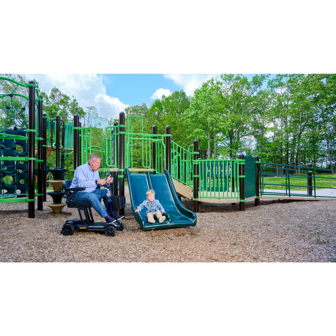 An adult riding a Golden Buzzaround GB220 CarryOn HD Travel Scooter cheers as a child slides at an outdoor playground framed by green equipment and trees, highlighting fun for all ages and abilities.