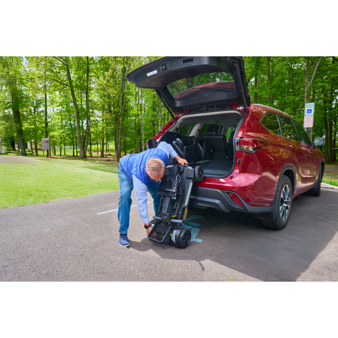 A person unloads a Golden Buzzaround GB220 CarryOn HD Travel Scooter from the trunk of a red SUV parked in a lot with green trees.