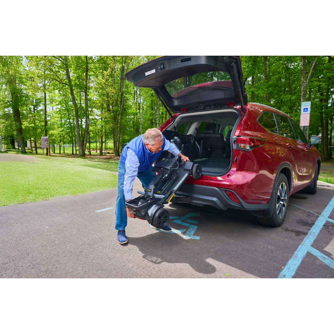 An older man unloads a Golden Buzzaround GB220 CarryOn HD Travel Scooter with a powerful brushless motor from the trunk of a red SUV parked in an accessible spot near woods.