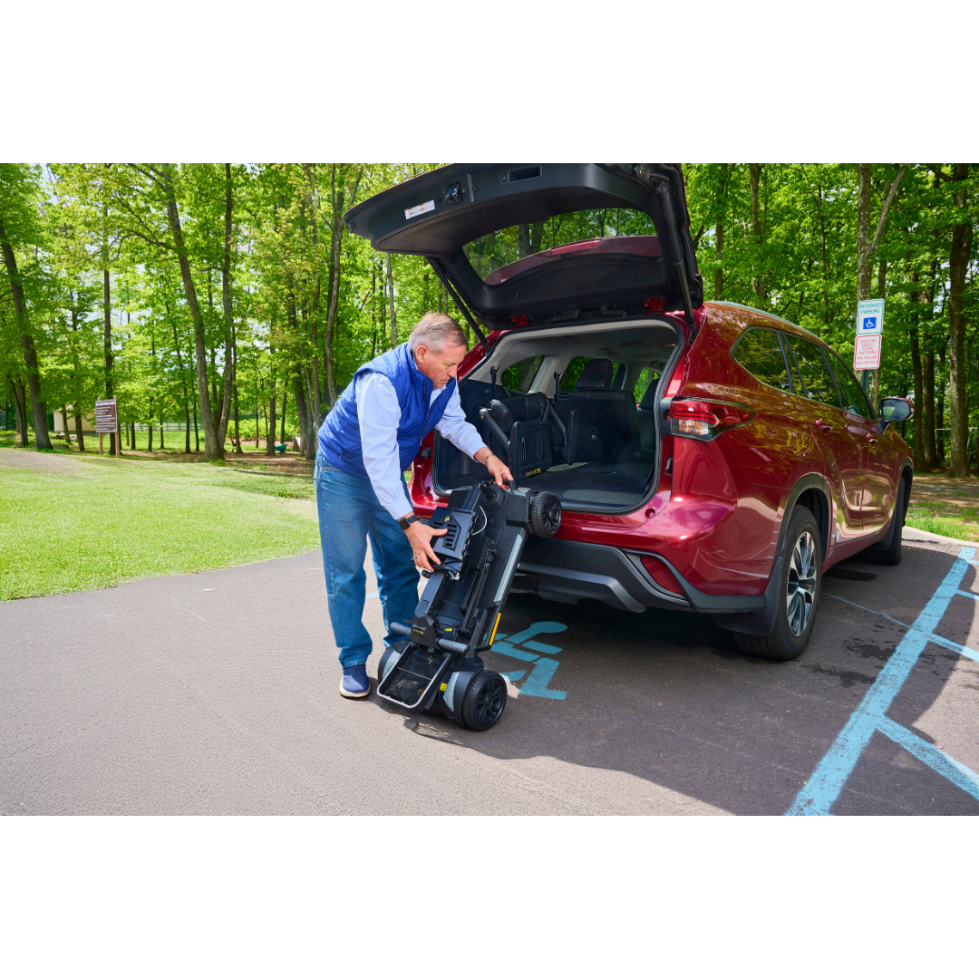 A man loads a Golden Buzzaround GB220 CarryOn HD Travel Scooter into the back of a red SUV parked in an accessible spot at a wooded park. The SUV’s rear hatch is open under sunny, clear skies.