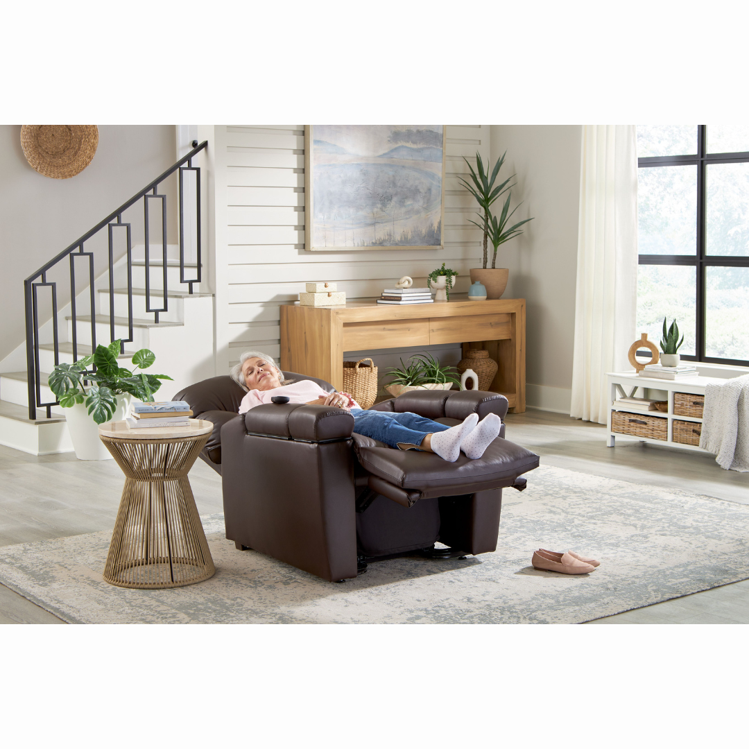 A person relaxing in a Golden Cloud+ PR511 Recliner with Lift Assist - Medium Wide, covered by a light blanket in a modern living room featuring a wooden side table, plants, a rug, stairs, and a large window with curtains.