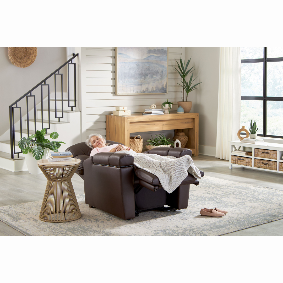 A person peacefully sleeps on a Golden Cloud+ PR511 Recliner with Lift Assist - Medium Wide, draped in a light blanket. The modern room showcases a staircase, wooden console table, plants, landscape painting, large windows, and pink slippers near the chair. The recliners AutoDrive adds to its 4-Zone Comfort Zone relaxation.